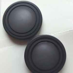 Peut inclure: Deux boutons en plastique noir avec un centre surélevé et trois cercles concentriques autour du bord. Les boutons sont sur un fond blanc avec le texte "boutons à la mode" et le numéro "146".