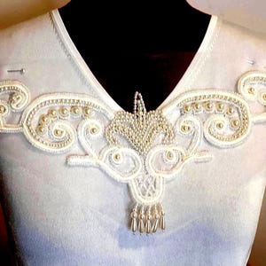 May include: White V-neck top with an elaborate embroidered design. The embroidery includes swirling patterns, small pearls, and a central beaded element with dangling teardrop beads. The top is displayed on a black mannequin.