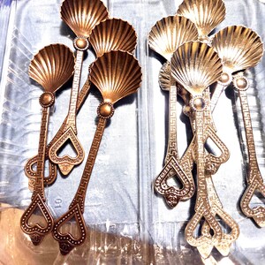 May include: A collection of metal spoons featuring shell-shaped bowls and long handles with a Greek key pattern. The handles terminate in a heart-shaped cutout design with a small circular logo emblem at the base of the bowl and the handle tip.