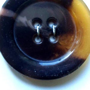 May include: An oval button with a tortoiseshell design in brown and yellow hues. The button features two sewing holes and is 38 mm in diameter. The number "703534U" is printed on the bottom.