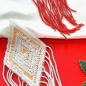 May include: Two diamond-shaped beaded patches featuring long fringe. One patch is entirely red with dense beadwork and sequins. The second patch is white with gold sequin accents and white fringe. Both items feature intricate hand-stitched beadwork.