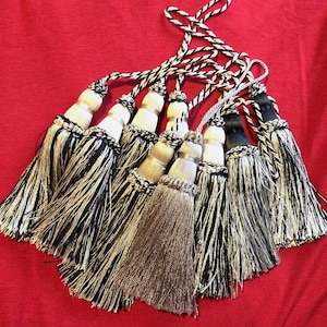 May include: A selection of decorative tassels in black, white, and silver. Each tassel features a cylindrical top with silver detailing, connected to a twisted cord. The background is a solid red.