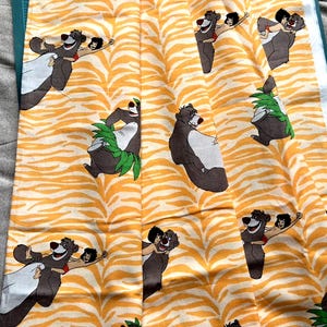 May include: A patterned fabric with a repeating design of a cartoon bear and a child, set against a yellow and white zebra stripe background. The bear is brown and white, and the child is wearing red. The fabric is likely cotton or a similar material.