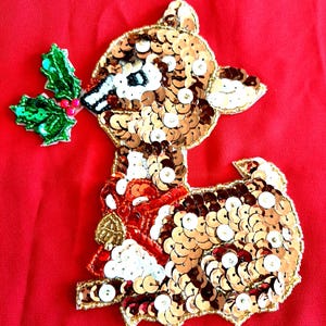 May include: A sequined reindeer applique with a red bow and a sprig of holly in its mouth, set against a red background. The reindeer is made of gold and brown sequins, with white and red accents. A festive holiday decoration.