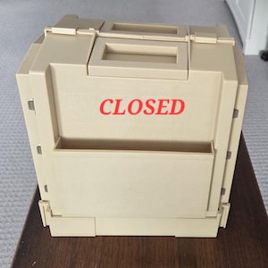 May include: A beige plastic storage case with a rectangular handle on top and a front pocket. The word CLOSED is printed in red capital letters on the front panel. The case features a structured design with visible metal hinges on the sides.
