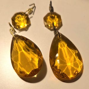 Peut inclure: Deux cristaux de lustre de couleur ambre, chacun composé d'une pièce supérieure octogonale à facettes reliée par un crochet métallique à un pendentif plus grand en forme de goutte à facettes. Les éléments en verre présentent une teinte jaune doré translucide avec des facettes géométriques qui captent et reflètent la lumière, créant un aspect brillant et chaleureux.