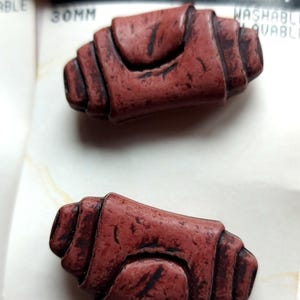 Peut inclure: Deux boutons rectangulaires brun rougeâtre avec une finition texturée imitation bois et des bords rainurés en couches. Chaque bouton présente un panneau central surélevé et mesure 30 mm, comme indiqué sur l'emballage. Les boutons sont présentés sur une carte blanche avec un code-barres et une pièce de monnaie canadienne visible sur le bord supérieur.
