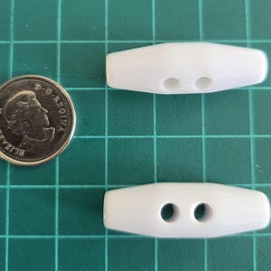 May include: Two white toggle buttons with two centered holes each, positioned next to a Canadian one-dollar coin for scale. The buttons have a smooth, elongated, and tapered shape, providing a clean and simple look for various fastening needs.
