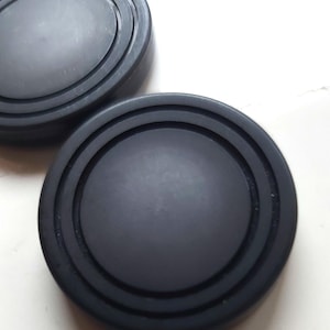 May include: Two black, circular objects with concentric grooves. One is oval-shaped, the other is round. The objects are made of a matte material and are set against a white background. The objects appear to be lids or stoppers.