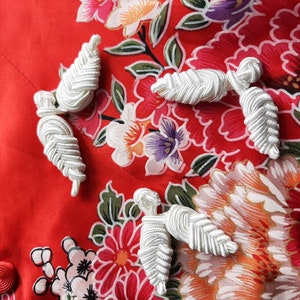 May include: Close-up of a red fabric with a floral pattern featuring large pink and white flowers, green leaves, and purple accents. White, decorative corded closures are sewn onto the fabric.