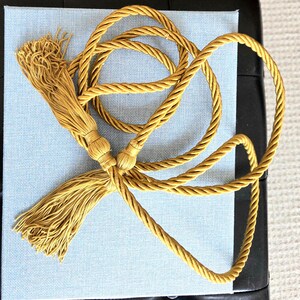 May include: Mustard yellow decorative tassel with a twisted rope design. The tassel is made of a soft, silky material and is attached to a matching rope. The rope is looped and knotted, creating an elegant accent piece. The tassel is lying on a light blue surface.