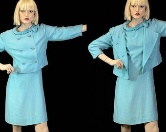May include: A light blue skirt suit with a double-breasted jacket and a bow detail at the neckline.