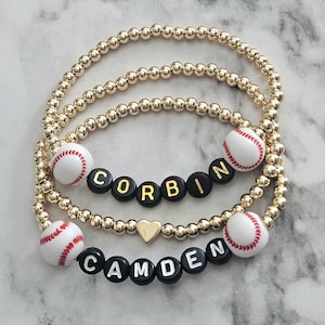 May include: Three gold beaded bracelets. Two bracelets have baseball-themed beads and the names "Corbin" and "Camden" spelled out in black letter beads. One bracelet has a gold heart charm.