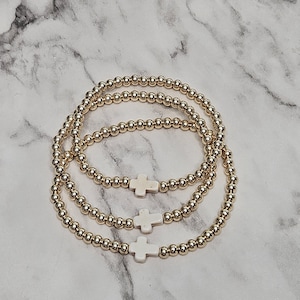 May include: Three gold beaded bracelets with white cross charms. The bracelets are arranged in a layered style, with each bracelet featuring small, round gold beads. The white cross charms are centered on each bracelet.