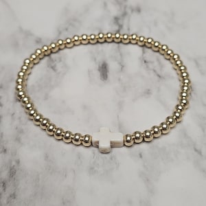 May include: Gold beaded bracelet with a white cross charm. The bracelet is made of small, round gold beads, with a smooth, polished finish. A white cross is centered on the bracelet, adding a touch of elegance and faith. The bracelet is approximately 18 cm in circumference.