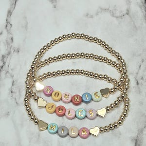 Op de afbeelding: Drie gouden kralenarmbanden met kleurrijke letterkralen met de namen "NONNIE", "JULES" en "MILA". Elke armband heeft een gouden hartbedel. De armbanden worden gepresenteerd op een wit-grijs marmeren oppervlak.
