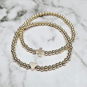 May include: Two gold beaded bracelets with a heart charm and a cross charm. The bracelets are stacked on top of each other.