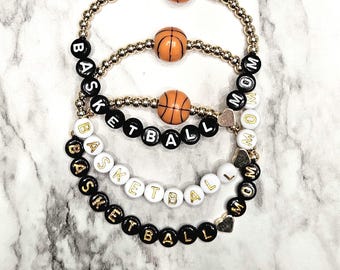 May include: Three stretch bracelets featuring gold-toned metallic beads and orange basketball-shaped beads. Two bracelets include black letter beads spelling BASKETBALL MOM, while one white bead bracelet spells BASKETBALL MOM with gold lettering.
