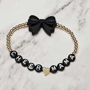 May include: A beaded bracelet featuring small metallic gold beads, black letter beads spelling CHEER MAMA, a central gold heart-shaped bead, and a prominent black bow-shaped bead at the top. The design uses a circular elastic cord construction.