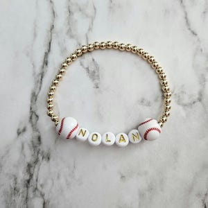 May include: A gold beaded bracelet with the name "NOLAN" spelled out in white letter beads with gold lettering. Two baseball beads are on the bracelet. The baseballs are white with red stitching.