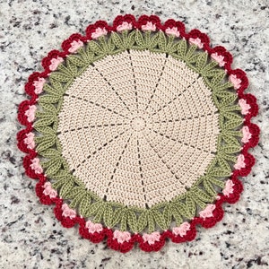 May include: A crocheted doily with a beige center and a green and pink border. The border features a repeating pattern of leaves and flowers.