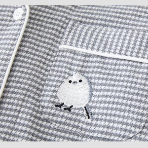 May include: Close-up of a grey and white houndstooth pajama top with white piping and buttons. A small pocket features an embroidered white bird with black details. The pajama top is made of a soft, textured fabric.