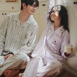 以下が含まれることがあります： 長袖のボタンダウンパジャマ2セット。ノッチドラペルと胸ポケット付き。1セットはライトグレーと白の縦縞模様、もう1セットはライトラベンダー地に白のストライプ柄。どちらも小さな刺繍グラフィックが施されています。