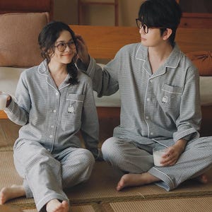 May include: Matching grey and white striped pyjama sets. Each set includes a button-down shirt with a small pocket and a pair of trousers. The shirts have a small embroidered design on the pocket. The pyjamas are worn by two people.