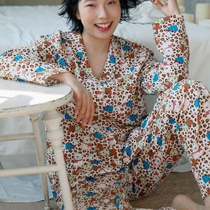 May include: A beige pajama set with a repeating pattern of cartoon bears, blue accents, and leopard print spots. The long-sleeved top has a collar and buttons. The matching pants are also patterned. The person is sitting on a woven mat.