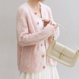 May include: A pale pink cardigan with a round neck and pink buttons. The cardigan is worn with a white dress and a cream-coloured handbag with a gold buckle. The handbag has a crocodile skin texture.