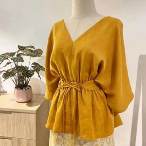 May include: Mustard yellow blouse with a V-neck and a tie-waist detail. The blouse has three-quarter sleeves and is paired with a yellow floral print skirt. The outfit is displayed on a mannequin.
