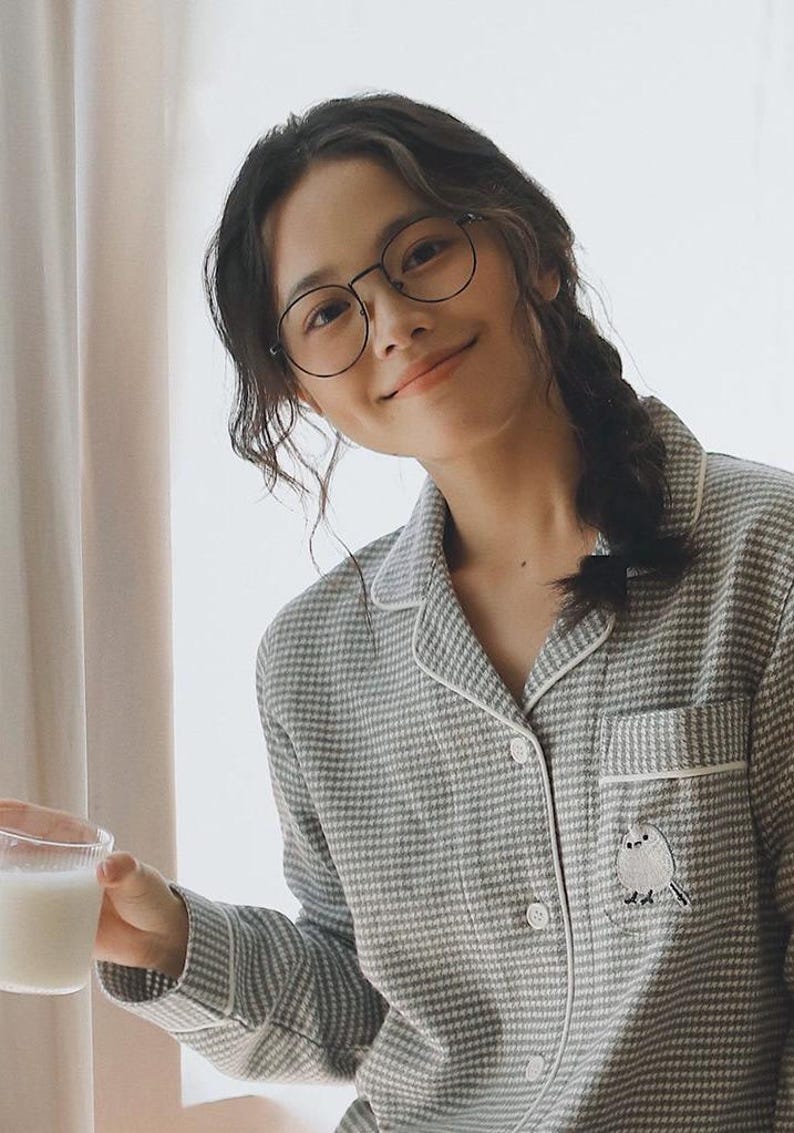 May include: A person wearing round glasses and a grey and white checked pajama set. The pajama top has a small bird embroidery on the pocket. The person is holding a glass of milk.