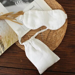 May include: A white linen sleep mask with an adjustable strap and a matching drawstring pouch. The mask and pouch are placed on a woven mat and a magazine. The sleep mask is designed to block out light for better sleep.