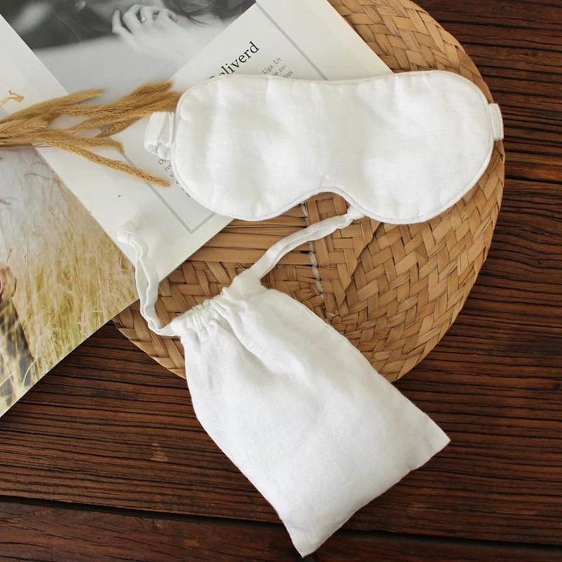 May include: A white linen sleep mask with an adjustable strap and a matching drawstring pouch. The mask and pouch are placed on a woven mat and a magazine. The sleep mask is designed to block out light for better sleep.