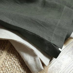 May include: Close-up of folded linen fabric in shades of gray and off-white. The fabric has a textured appearance and is layered, with the edges of the fabric visible. The background includes a woven rug and a wooden surface.