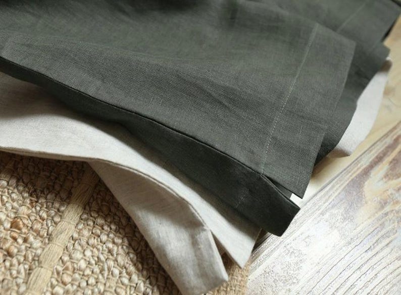 May include: Close-up of folded linen fabric in shades of gray and off-white. The fabric has a textured appearance and is layered, with the edges of the fabric visible. The background includes a woven rug and a wooden surface.