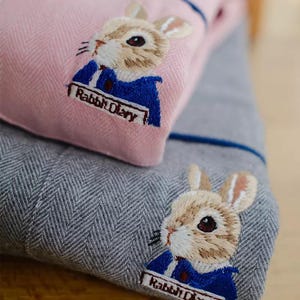 Puede incluir: Primer plano de dos piezas de tela, una rosa y otra gris, con un patrón de espiga. Cada pieza presenta un conejo bordado con una chaqueta azul. El texto "Rabbit Diary" está bordado debajo.