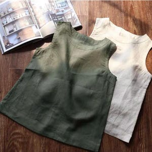 May include: Two sleeveless linen tops in olive green and off-white are displayed on a wooden surface. The tops have a simple, minimalist design with a rounded neckline. An open book with images of pottery and furniture is in the background.