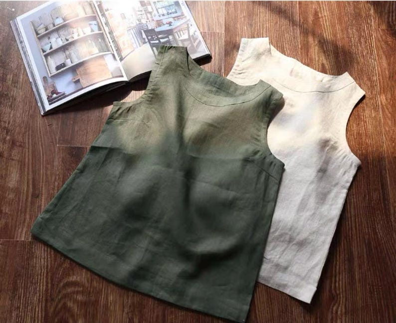 May include: Two sleeveless linen tops in olive green and off-white are displayed on a wooden surface. The tops have a simple, minimalist design with a rounded neckline. An open book with images of pottery and furniture is in the background.