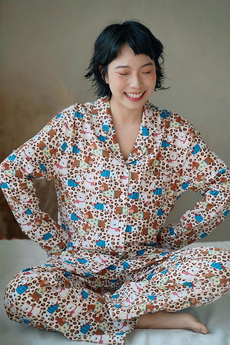 May include: A beige pajama set with a repeating pattern of cartoon bears and leopard spots. The long-sleeved top has a button-down front and a collar. The matching pants complete the set. The pajamas are made of a soft material.