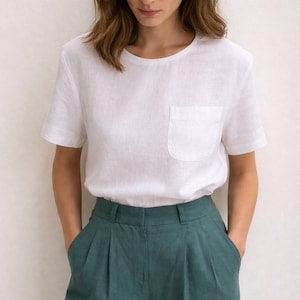 Puede incluir: Una persona con una camiseta de lino blanca con bolsillo en el pecho y pantalones plisados verde azulado. El atuendo es simple e informal, adecuado para el uso diario. La ropa parece estar hecha de fibras naturales.