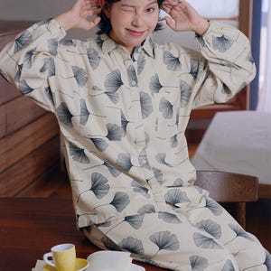 Op de afbeelding: Een tweedelige pyjamaset bestaande uit een overhemd met lange mouwen en knoopsluiting en een bijpassende broek van crèmekleurige stof. De set is voorzien van een herhalend patroon van donkerblauwe ginkgo-bladillustraties over het kledingstuk.