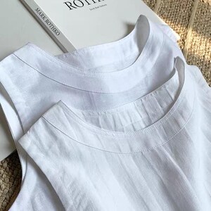 May include: Close-up of two white sleeveless tops with a round neckline. The tops are made of a textured fabric and appear to be linen or a similar material. The text "MARK ROTHKO" is visible on a book in the background.