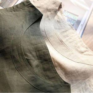 May include: Close-up of two linen shirts, one in a muted olive green and the other in a natural beige. The shirts have a simple, scoop neckline and are made of a textured fabric. The stitching detail is visible along the neckline.