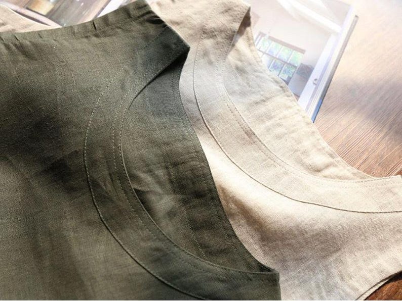 May include: Close-up of two linen shirts, one in a muted olive green and the other in a natural beige. The shirts have a simple, scoop neckline and are made of a textured fabric. The stitching detail is visible along the neckline.