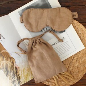 May include: A tan-colored sleep mask and matching drawstring bag are displayed on a magazine. The sleep mask has an elastic band. The bag is closed with a drawstring. The items are made of a soft, textured fabric. The background includes a woven mat.