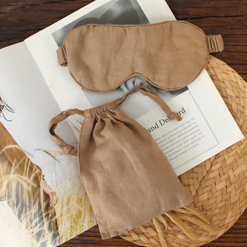 May include: A tan-colored sleep mask and matching drawstring bag are displayed on a magazine. The sleep mask has an elastic band. The bag is closed with a drawstring. The items are made of a soft, textured fabric. The background includes a woven mat.