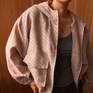May include: A light pink jacket with a hood and a zipper closure. The jacket has a striped pattern and two front pockets. The sleeves are long and gathered at the wrists. The jacket is open, revealing a gray tank top underneath.