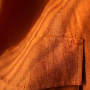 May include: Close-up of a textured, orange fabric with a small pocket. The fabric has a woven appearance and is illuminated with shadows, creating a play of light and dark. The pocket is rectangular and stitched onto the fabric.