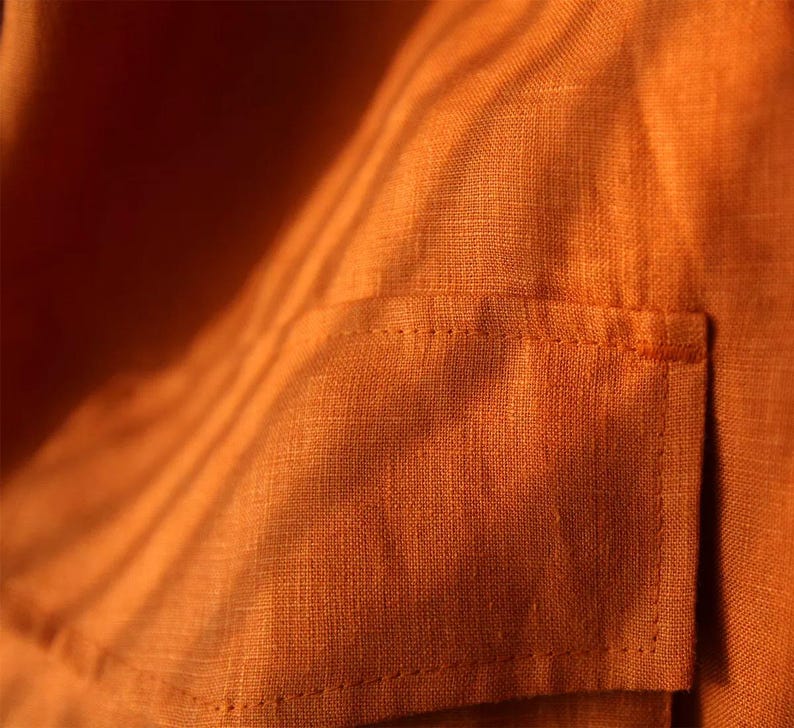 May include: Close-up of a textured, orange fabric with a small pocket. The fabric has a woven appearance and is illuminated with shadows, creating a play of light and dark. The pocket is rectangular and stitched onto the fabric.